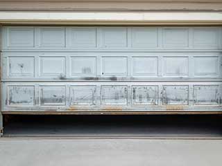 Damaged residential garage door stuck halfway open with visible dirt and rust.